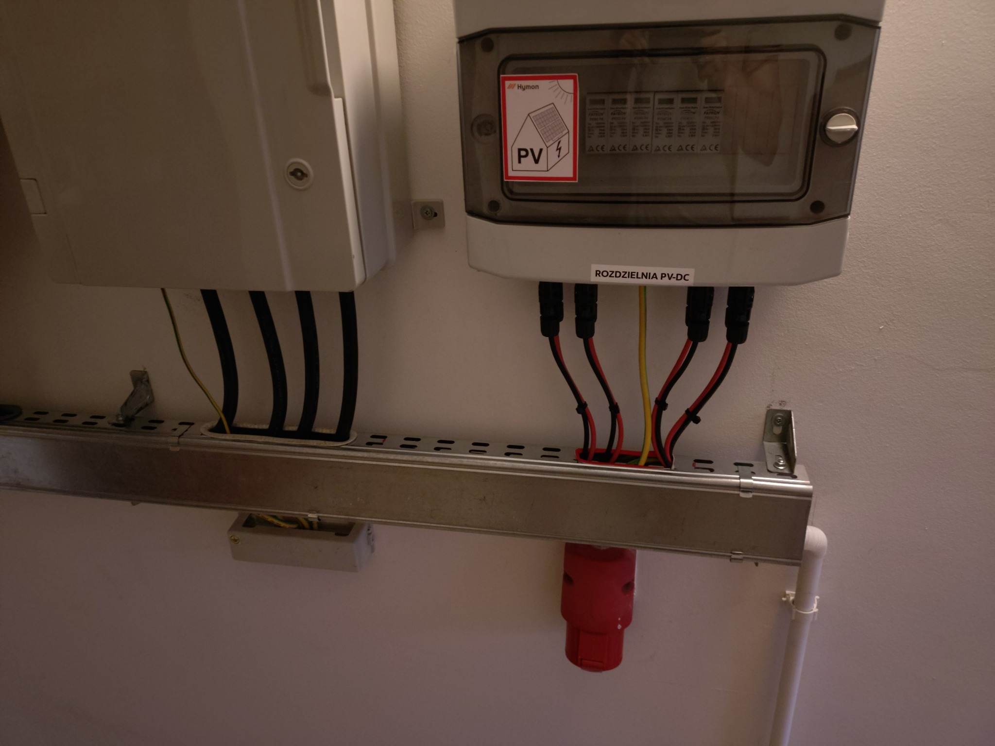 Rozdzielnia PV-DC z widocznymi przewodami i oznaczeniem PV, zamontowana na metalowej szynie przy białej ścianie. Widoczne elementy instalacji fotowoltaicznej.