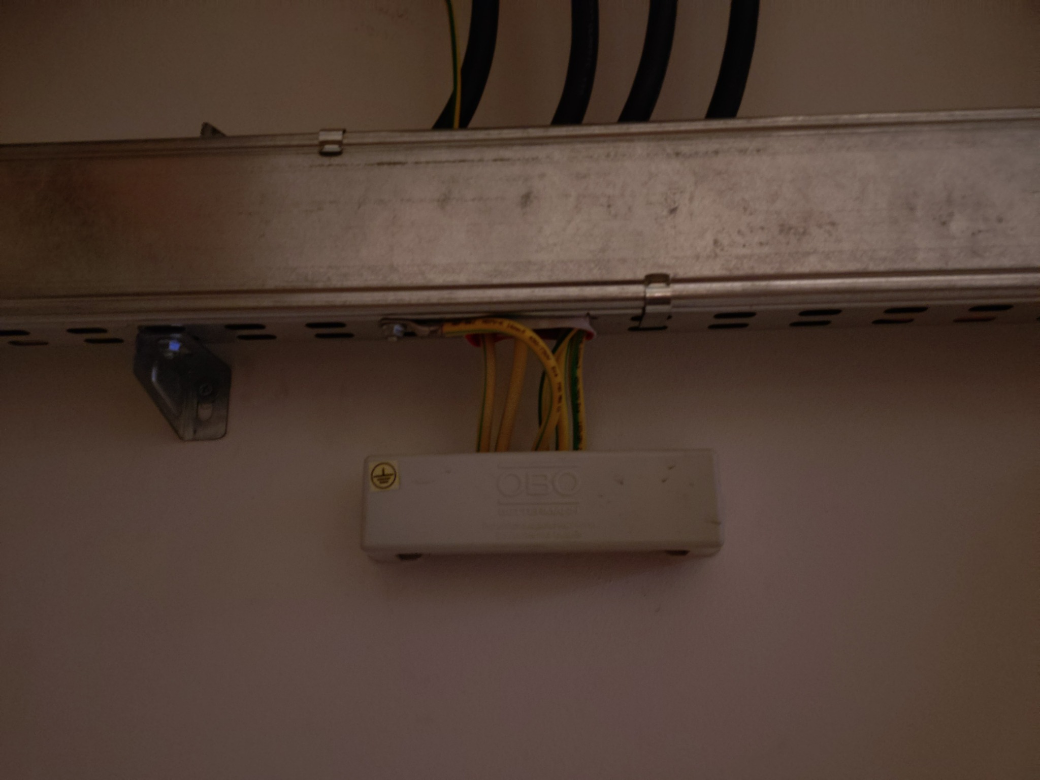 Metalowa szyna z przewodami elektrycznymi przymocowana do ściany, widoczna puszka instalacyjna z symbolem uziemienia. Detal instalacji.