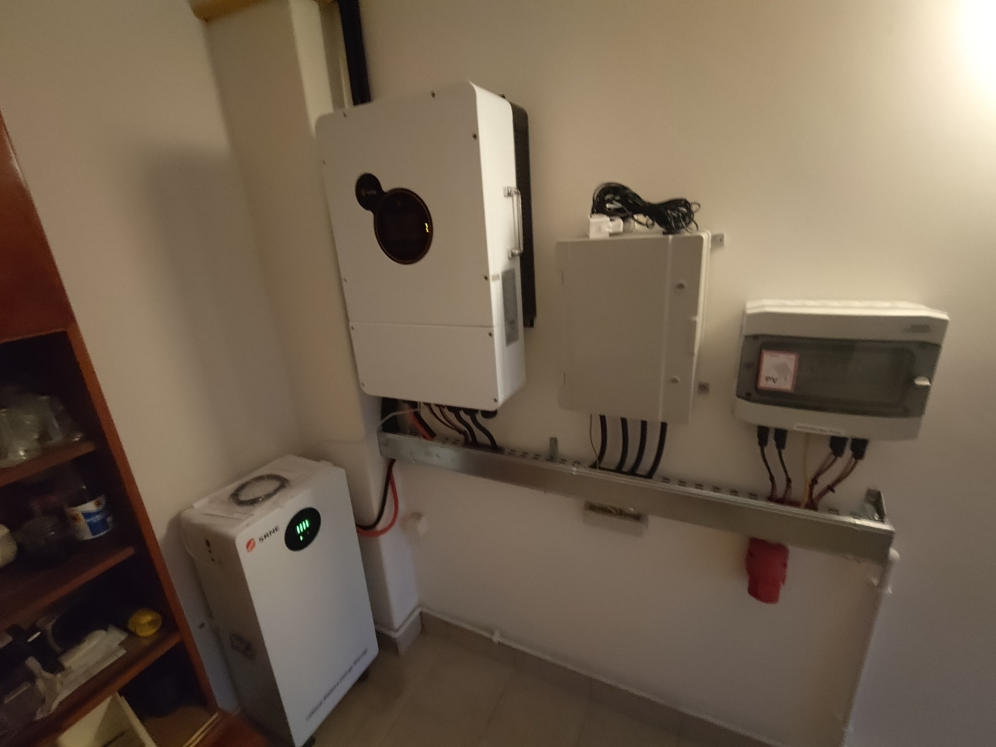 Biały inwerter fotowoltaiczny, akumulator energii i skrzynka bezpiecznikowa zamontowane na ścianie wewnątrz budynku. Widoczne kable i elementy instalacji.