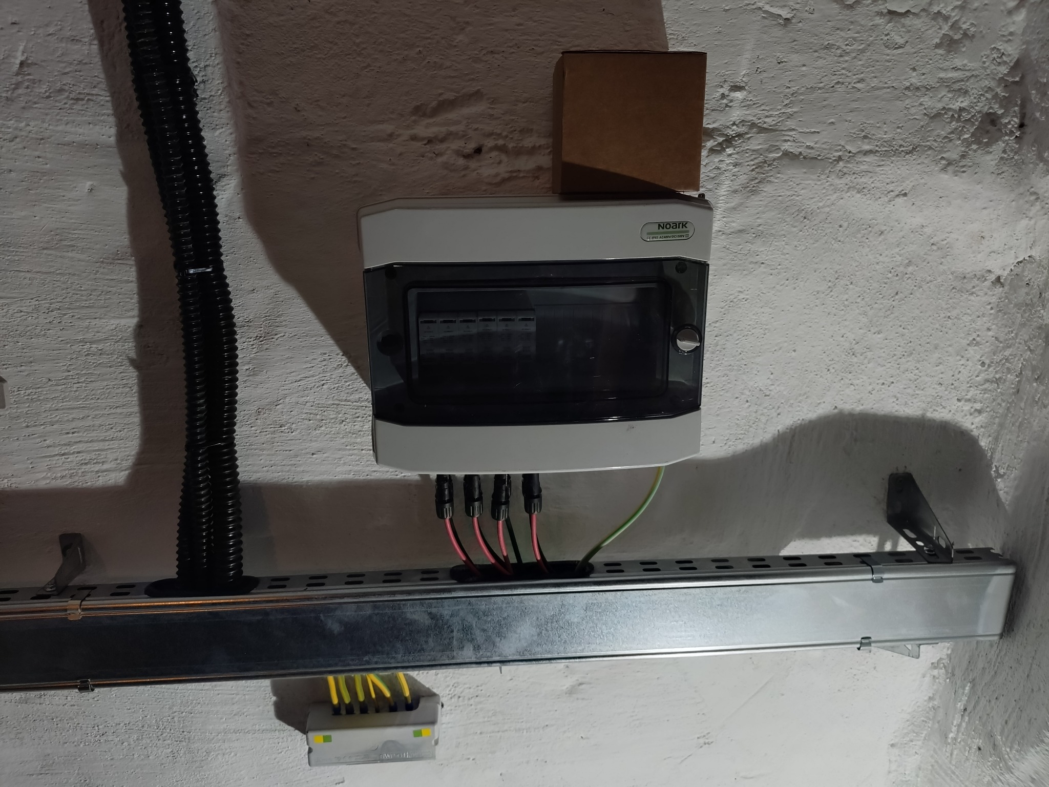 Szara skrzynka elektryczna Noark z podłączonymi przewodami na metalowej szynie montażowej, umieszczona na tle białej, chropowatej ściany. Widoczna czarna rura karbowana.