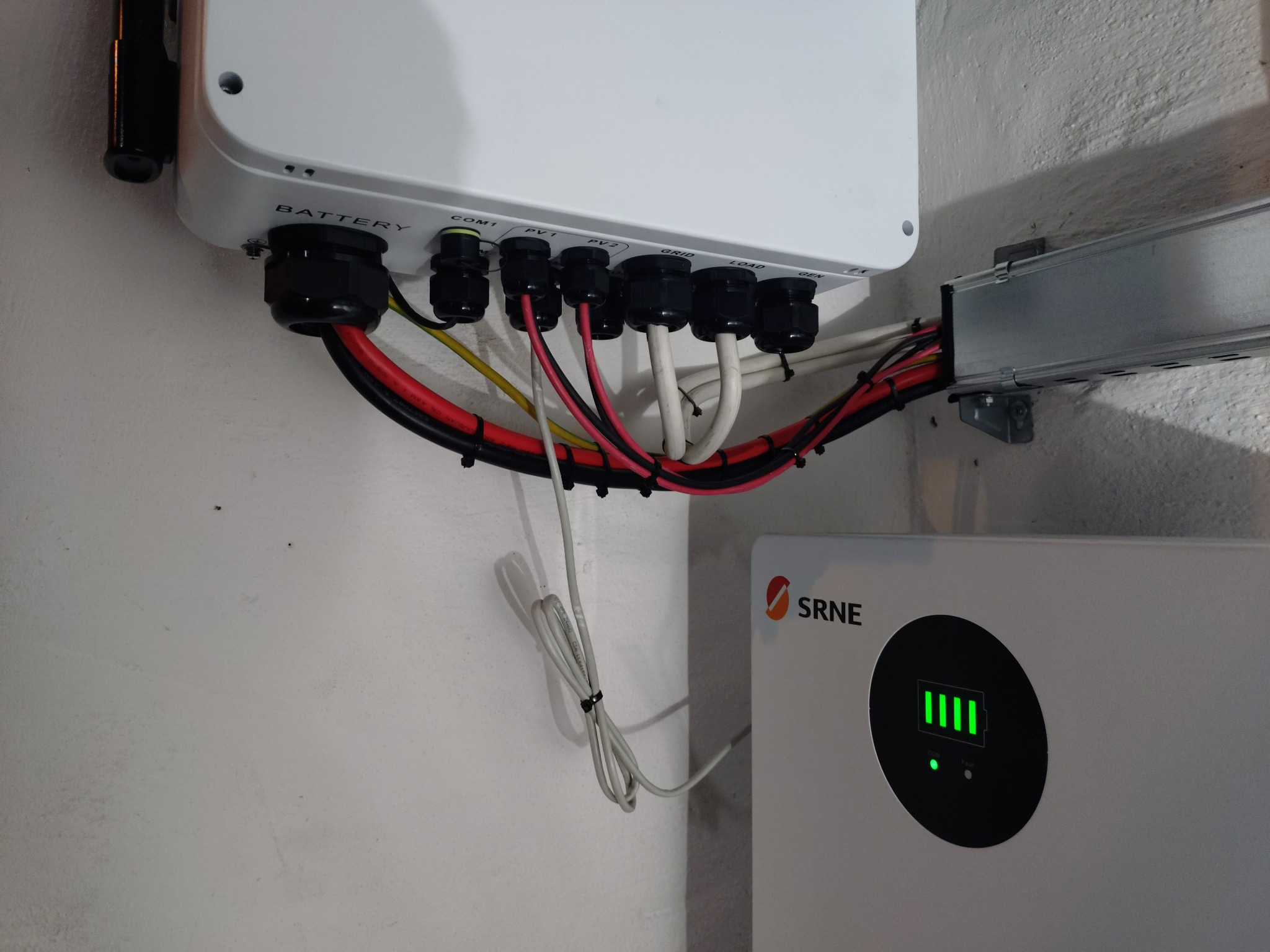 Podłączenie przewodów do falownika i magazynu energii SRNE. Widoczne kable w czarnych peszlach, listwa montażowa i wskaźnik naładowania baterii.