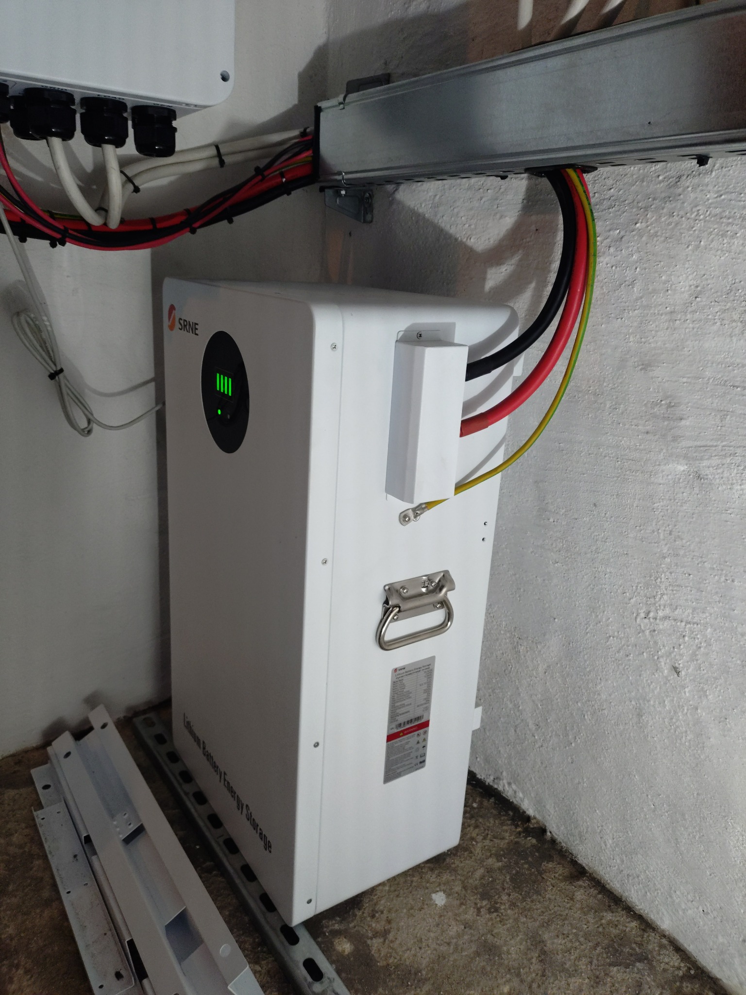 Biały magazyn energii SRNE z podłączonymi kablami, zamontowany w rogu pomieszczenia. Widoczne elementy montażowe i kable w peszlach.