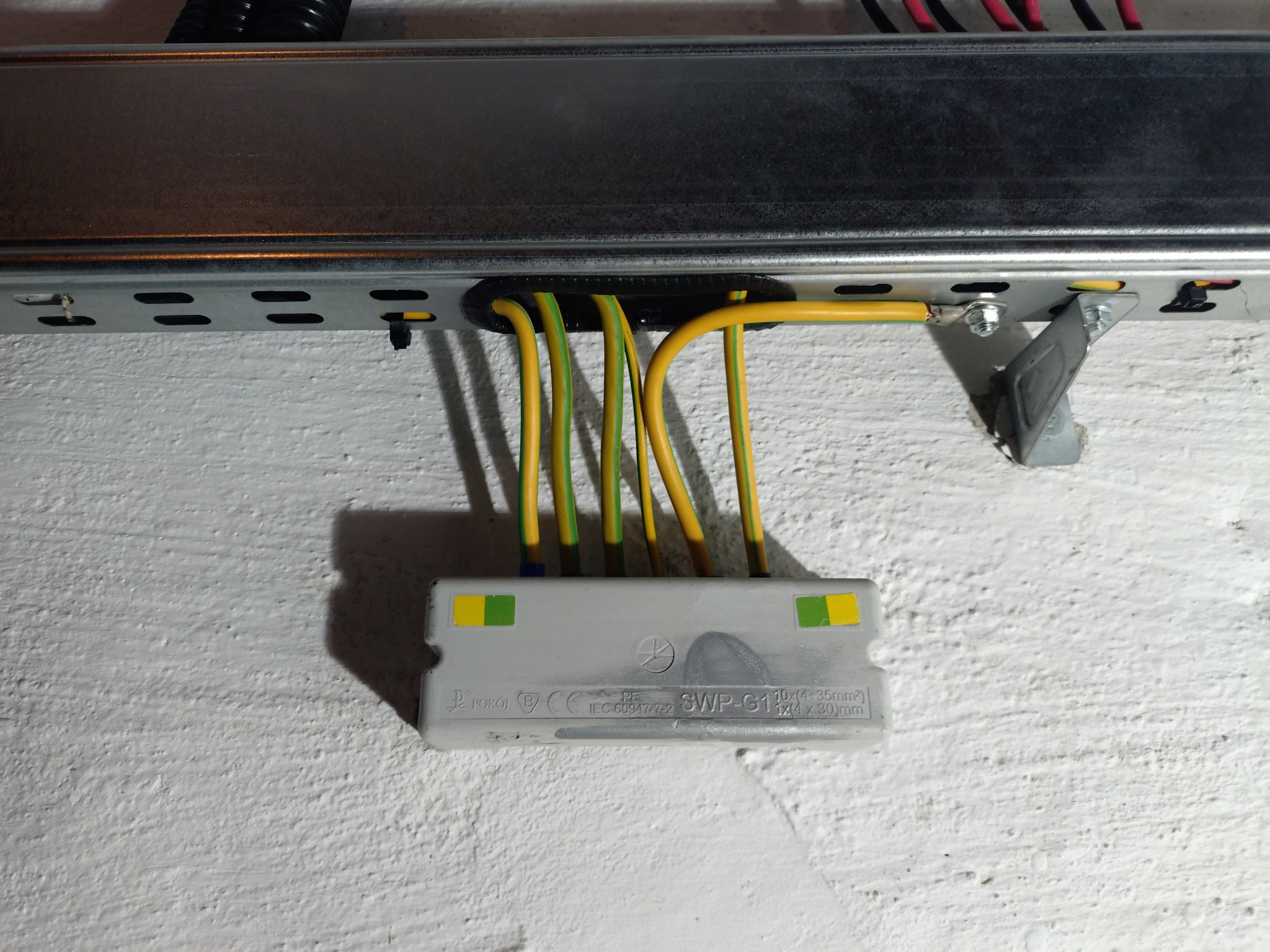 Szyna montażowa z podłączonymi przewodami elektrycznymi (żółto-zielone) i złączką. Widoczne oznaczenia na złączce. Biała ściana w tle.