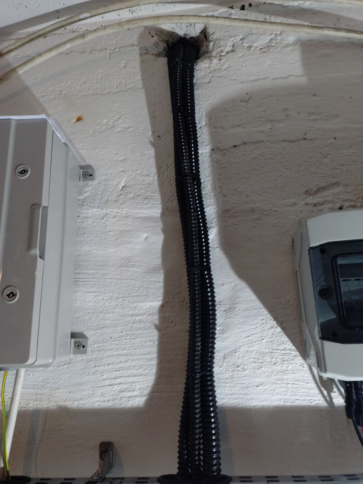 Kable w peszlu wchodzące do ściany, obok skrzynka elektryczna i licznik. Widoczne mocowania i fragment ściany z fakturą.