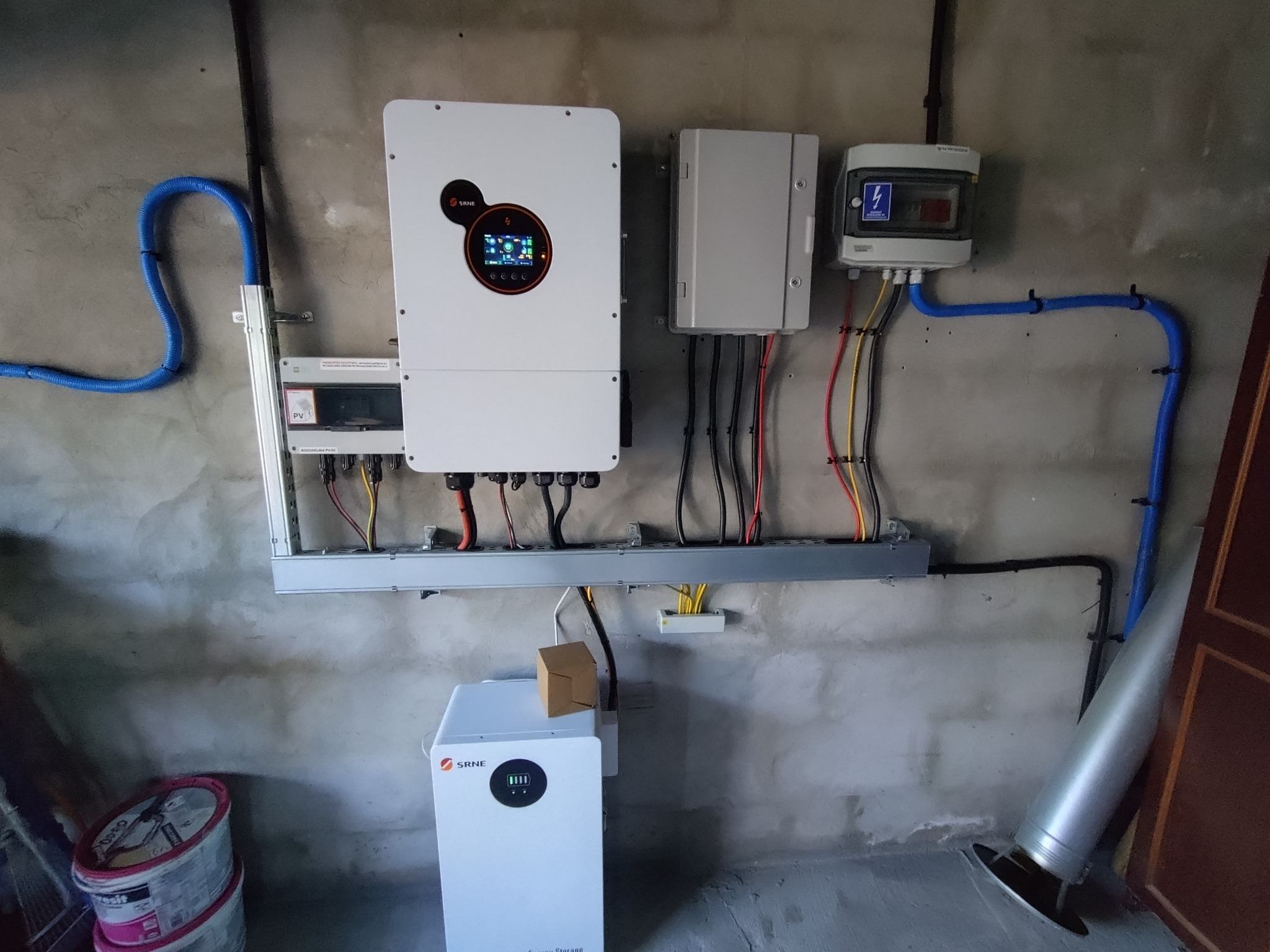 Kompletna instalacja fotowoltaiczna z inwerterem, skrzynką elektryczną i magazynem energii SRNE na szarej ścianie. Widoczne kable i rury instalacyjne.