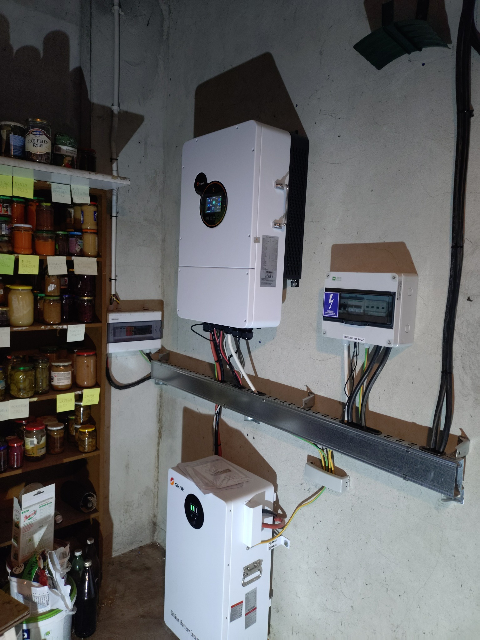 Kompletna instalacja fotowoltaiczna w piwnicy: inwerter, rozdzielnia PV-AC, magazyn energii SRNE, kable w korytku. Obok regał z przetworami.