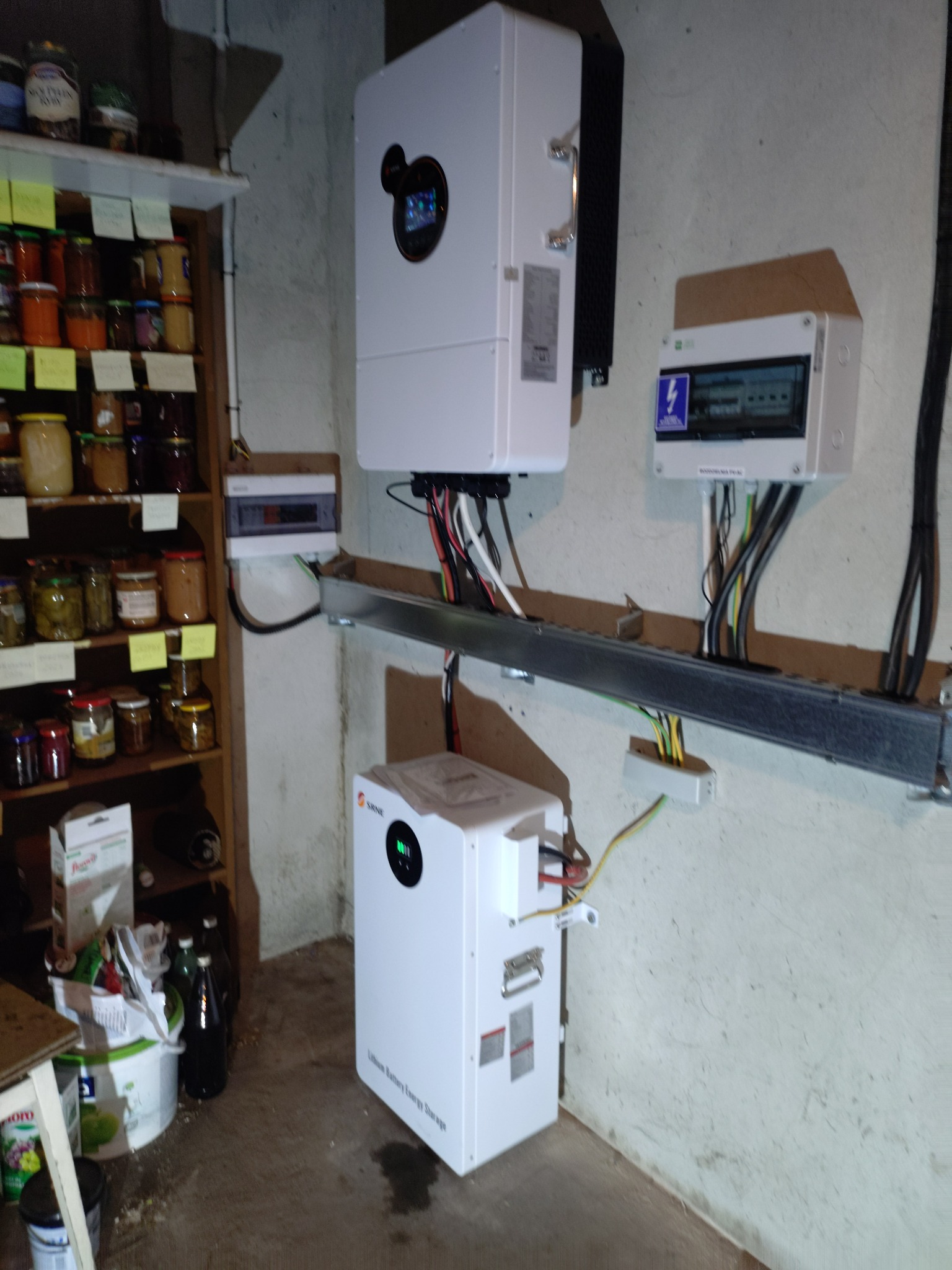 Biały inwerter fotowoltaiczny i akumulator energii zamontowane na ścianie w piwnicy, widoczne kable i skrzynka elektryczna. Obok regał z przetworami.