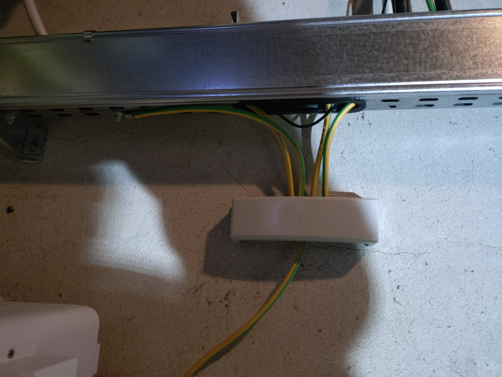 Szyna montażowa z przewodami ochronnymi (żółto-zielonymi) wchodzącymi do puszki elektrycznej na szorstkiej ścianie. Widoczne kable i elementy instalacji.