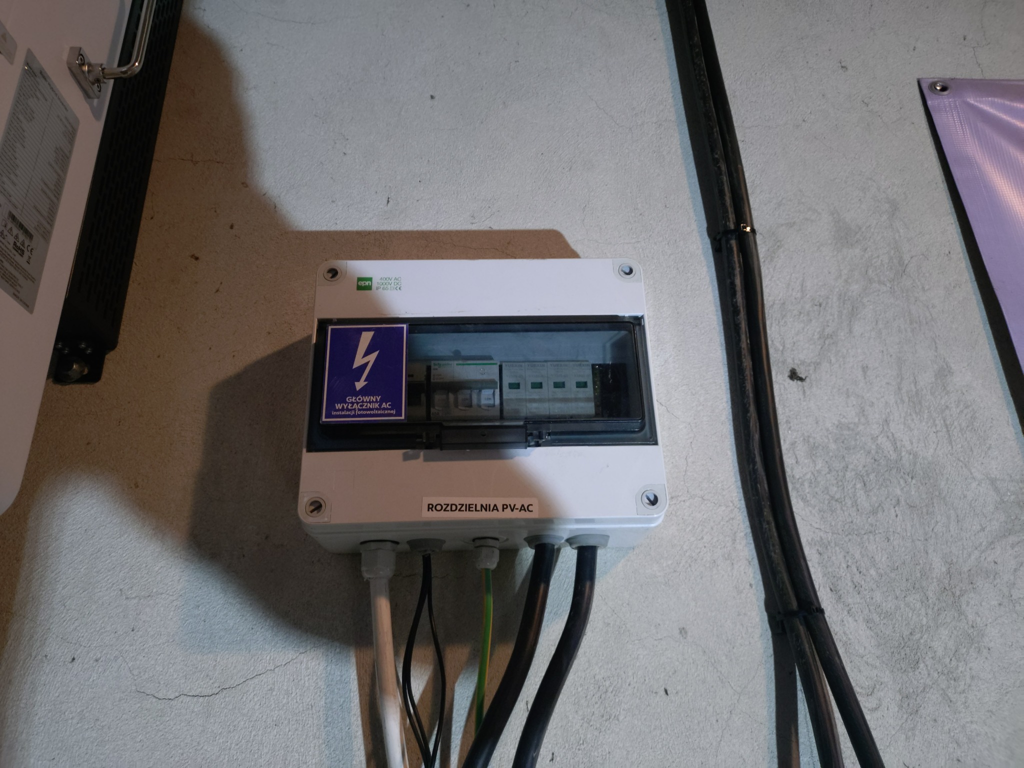 Rozdzielnia PV-AC z widocznymi przewodami i naklejką informacyjną o głównym wyłączniku AC instalacji fotowoltaicznej, zamontowana na szarej ścianie.