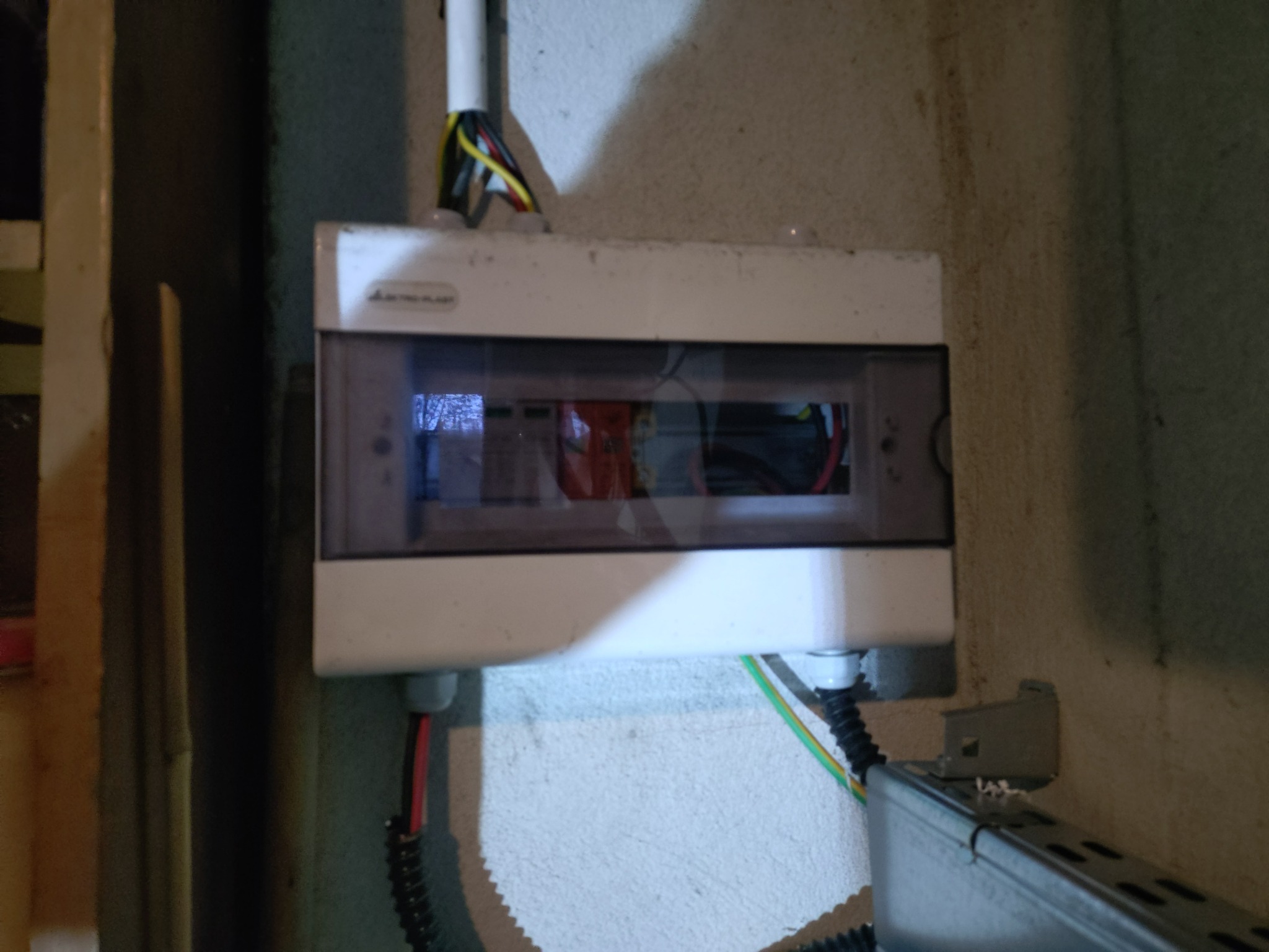 Zamknięta skrzynka elektryczna z widocznymi przewodami na szarej ścianie. Instalacja wykonana schludnie, z dbałością o detale. Widoczne kable w kolorach: żółty, czarny, czerwony.
