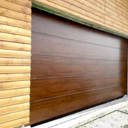 Brązowa, segmentowa brama garażowa zamontowana w drewnianym budynku z widocznym oświetleniem zewnętrznym i betonową wylewką u dołu.