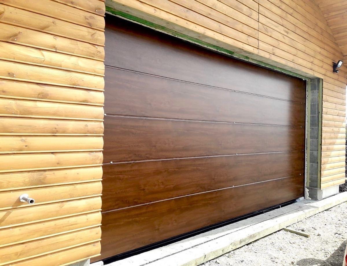 Brązowa, segmentowa brama garażowa zamontowana w drewnianym budynku z widocznym oświetleniem zewnętrznym i betonową wylewką u dołu.