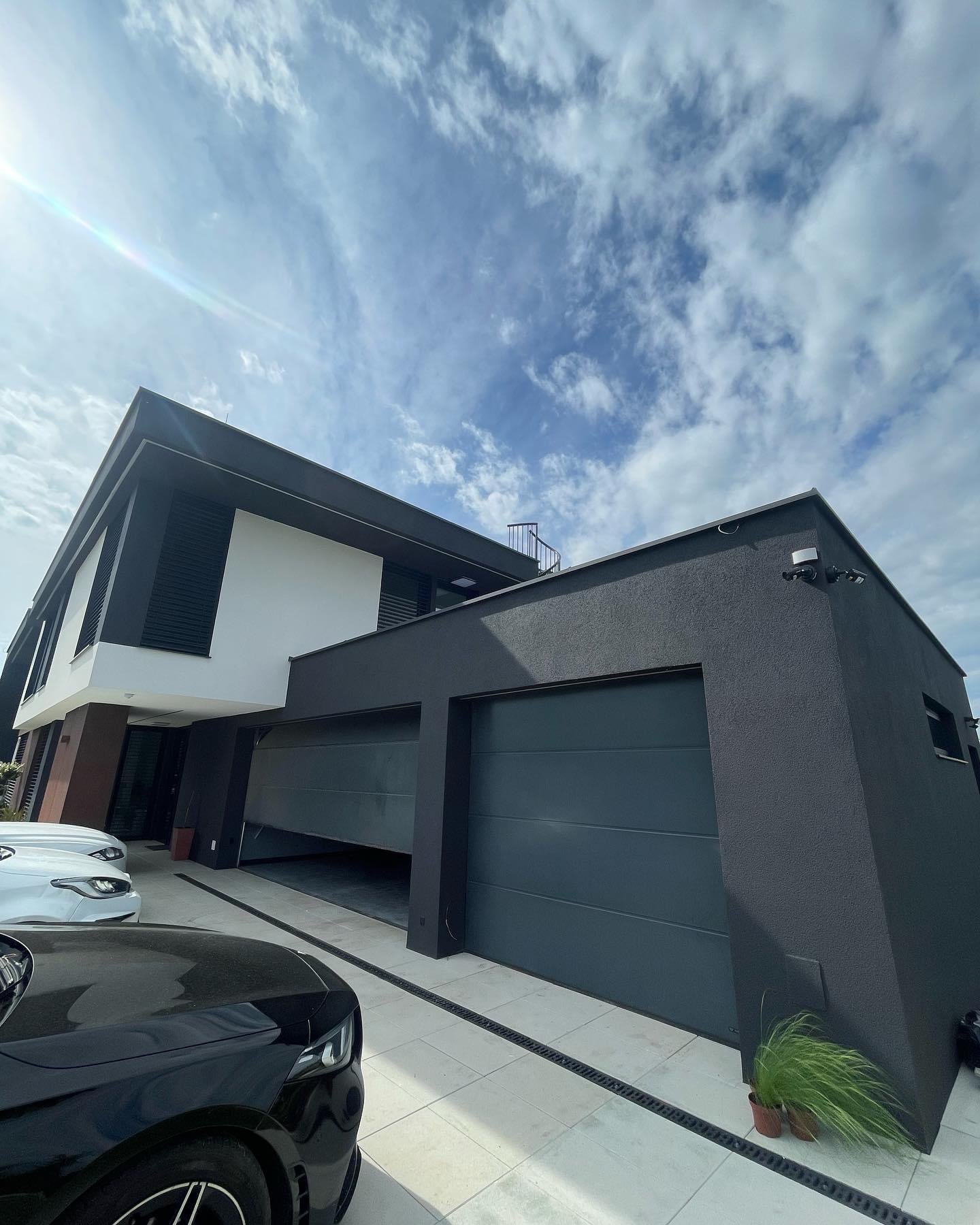Nowoczesny dom z grafitową elewacją, widoczne dwie bramy garażowe, jedna otwarta, oraz dwa samochody na podjeździe w słoneczny dzień.