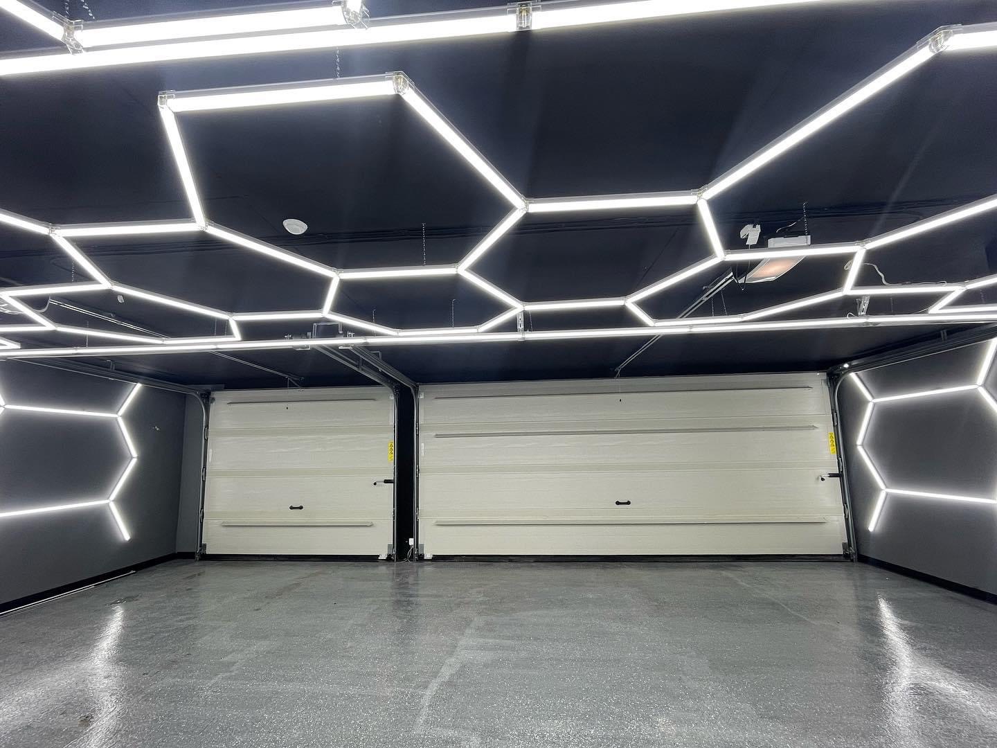 Wnętrze garażu z dwiema częściowo otwartymi, segmentowymi bramami garażowymi w kolorze złamanej bieli, oświetlone geometrycznymi lampami LED na suficie i ścianach.