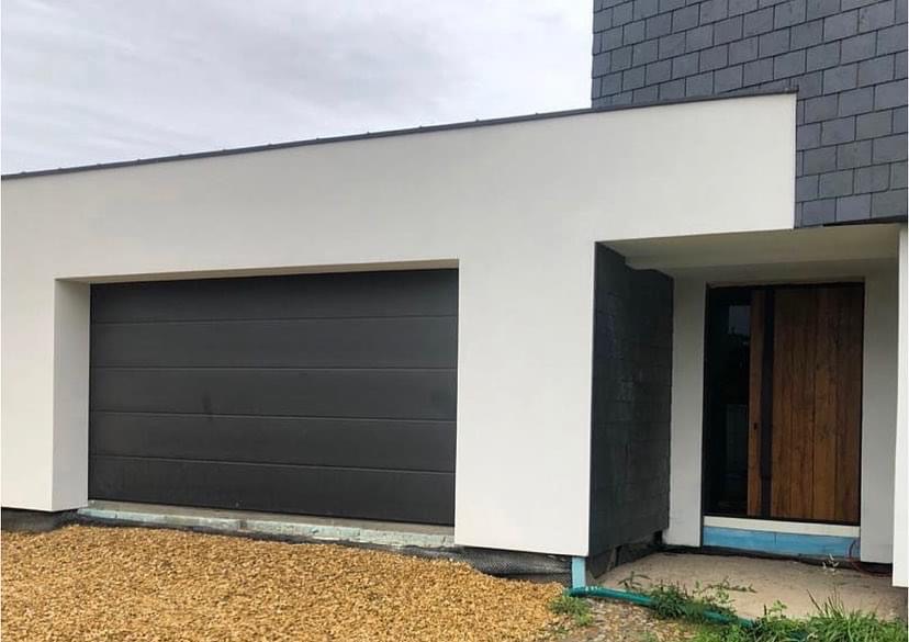 Nowoczesna brama garażowa segmentowa w kolorze antracytowym, zamontowana w białej bryle budynku jednorodzinnego z elementami szarej dachówki.