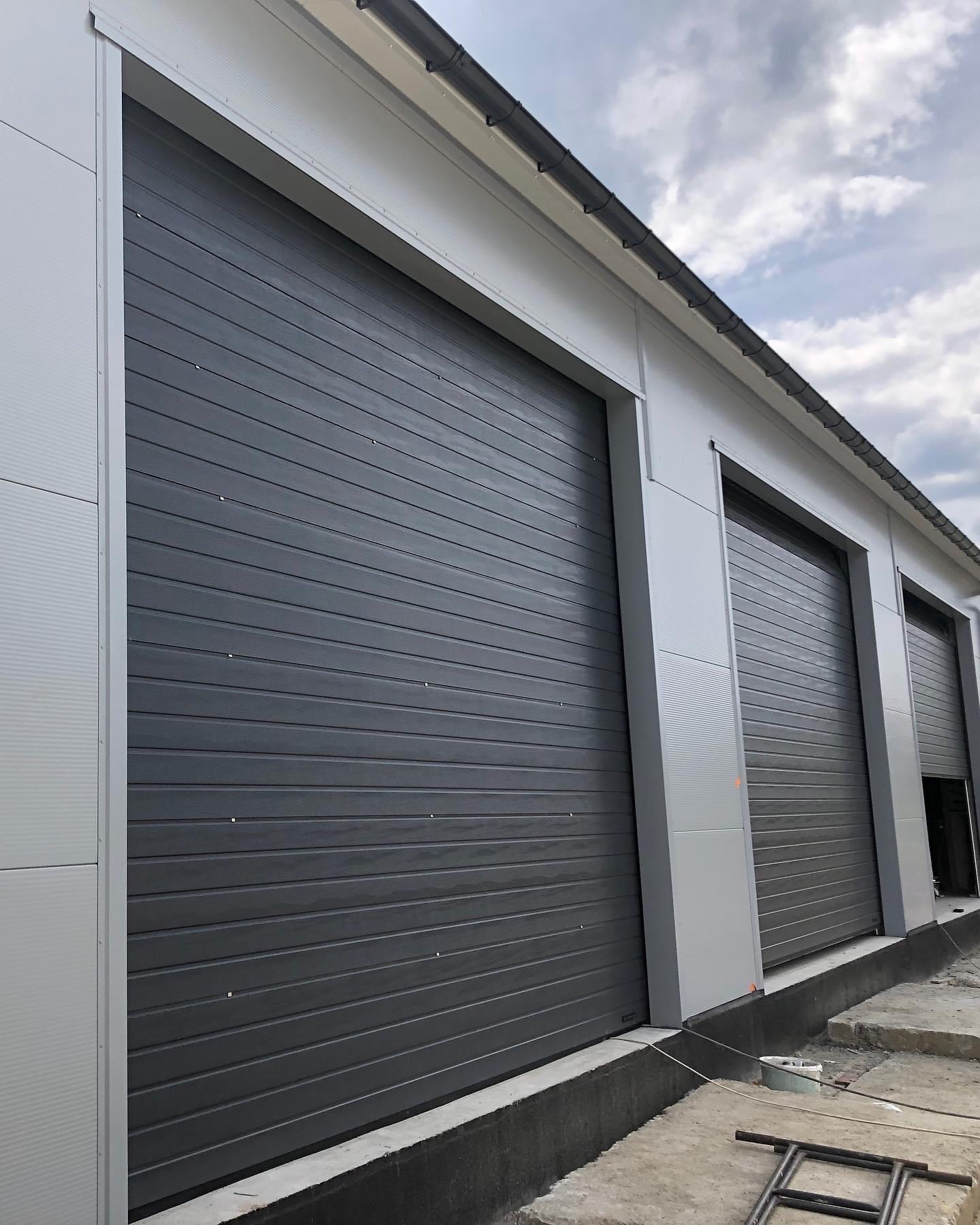 Zewnętrzny widok hali magazynowej z trzema zamkniętymi, segmentowymi bramami w kolorze antracytowym, widoczne elementy konstrukcyjne budynku i fragment drabiny na placu budowy.
