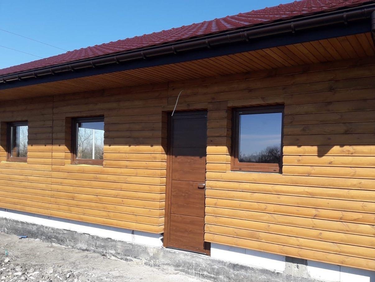 Dom z drewnianą elewacją, brązowymi oknami i drzwiami, oraz ciemnoczerwonym dachem, widziany w słoneczny dzień.