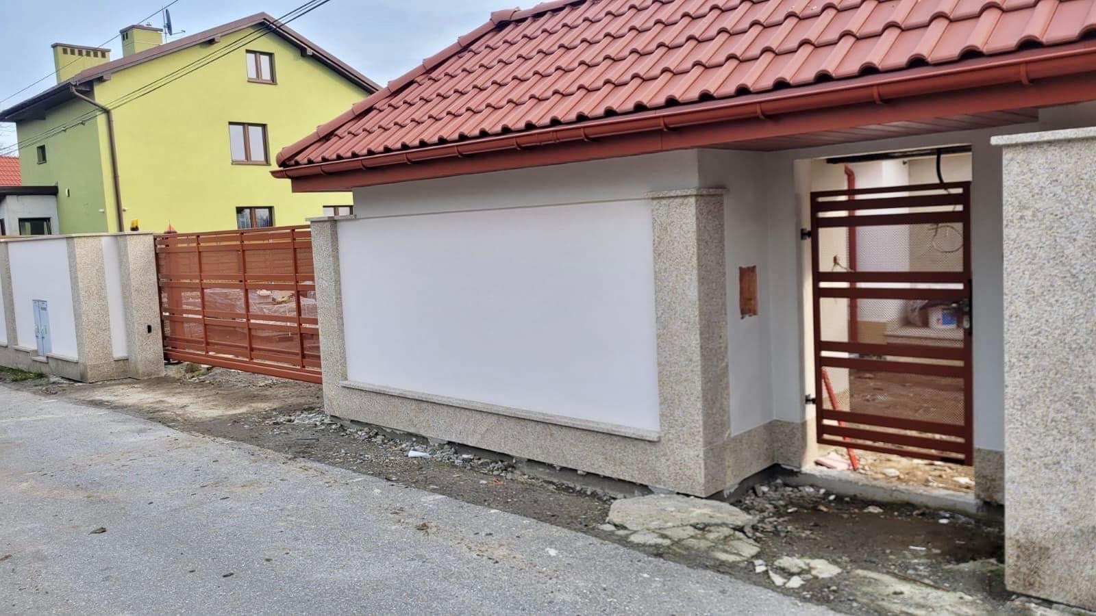 Nowoczesna brama wjazdowa przesuwna i furtka z poziomych profili w kolorze brązowym, zamontowane w ogrodzeniu z białymi panelami i elementami z jasnego granitu, na tle żółtego domu z czerwoną...