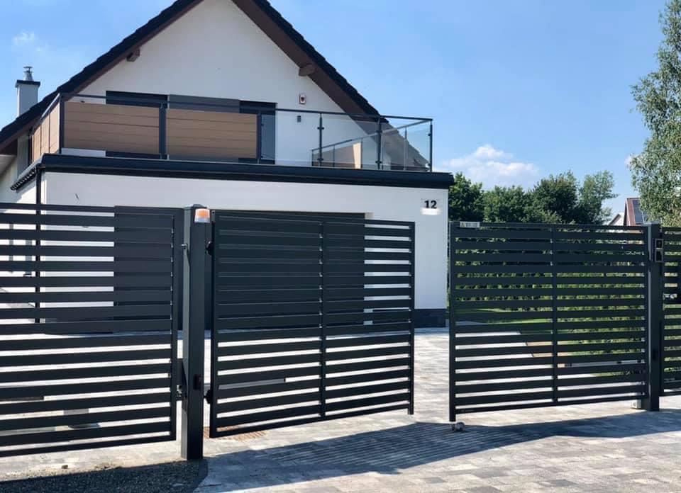 Nowoczesna, pozioma, metalowa brama wjazdowa w kolorze grafitowym, częściowo otwarta, z automatyką i lampą ostrzegawczą, na tle domu z balkonem z drewnianymi panelami i szklanymi barierkami.