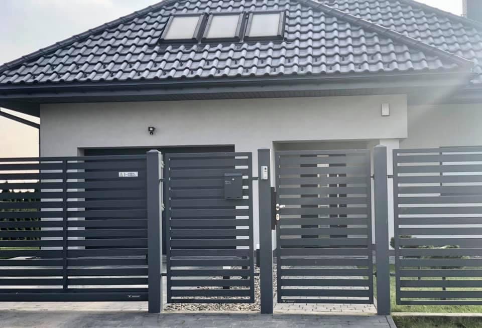 Nowoczesna, pozioma brama wjazdowa w kolorze grafitowym, z furtką i skrzynką na listy, na tle domu z ciemnoszarą dachówką i trzema oknami dachowymi.