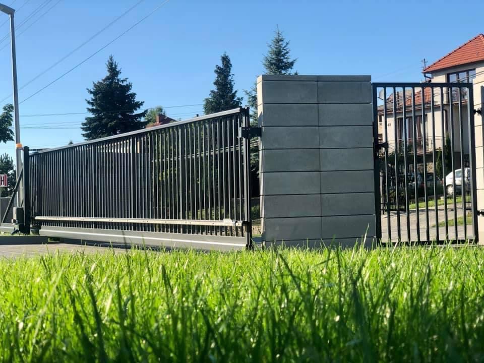 Nowoczesna brama wjazdowa przesuwna z metalowymi prętami i betonowym słupkiem, zainstalowana w Krakowie, widziana z perspektywy trawnika.