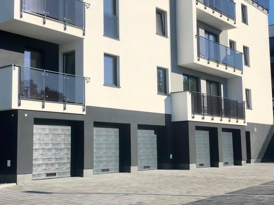 Elewacja nowoczesnego budynku wielorodzinnego z szarymi segmentowymi bramami garażowymi o poziomej fakturze drewna, balkonami z szklanymi balustradami i szarymi oknami.
