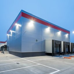 ART-BHP - Nowoczesny magazyn z rampami rozładunkowymi oświetlony lampami zewnętrznymi, widok z parkingu o zmierzchu. Czerwony pas na górze budynku.