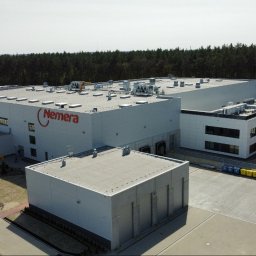 ART-BHP - Zdjęcie z lotu ptaka przedstawia budynek firmy Nemera z parkingiem, zielenią i lasem w tle. Widoczne kontenery na śmieci i samochody na parkingu. Jasne niebo.