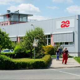 ART-BHP - Budynek firmy AE Group w słoneczny dzień. Pracownicy w odzieży roboczej wchodzą do budynku. Widoczne logo firmy na fasadzie i zadbana zieleń wokół.