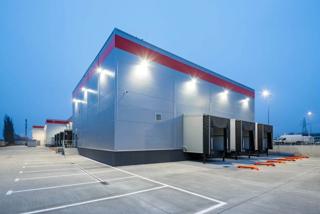 Nowoczesny magazyn z rampami rozładunkowymi oświetlony lampami zewnętrznymi, widok z parkingu o zmierzchu. Czerwony pas na górze budynku.