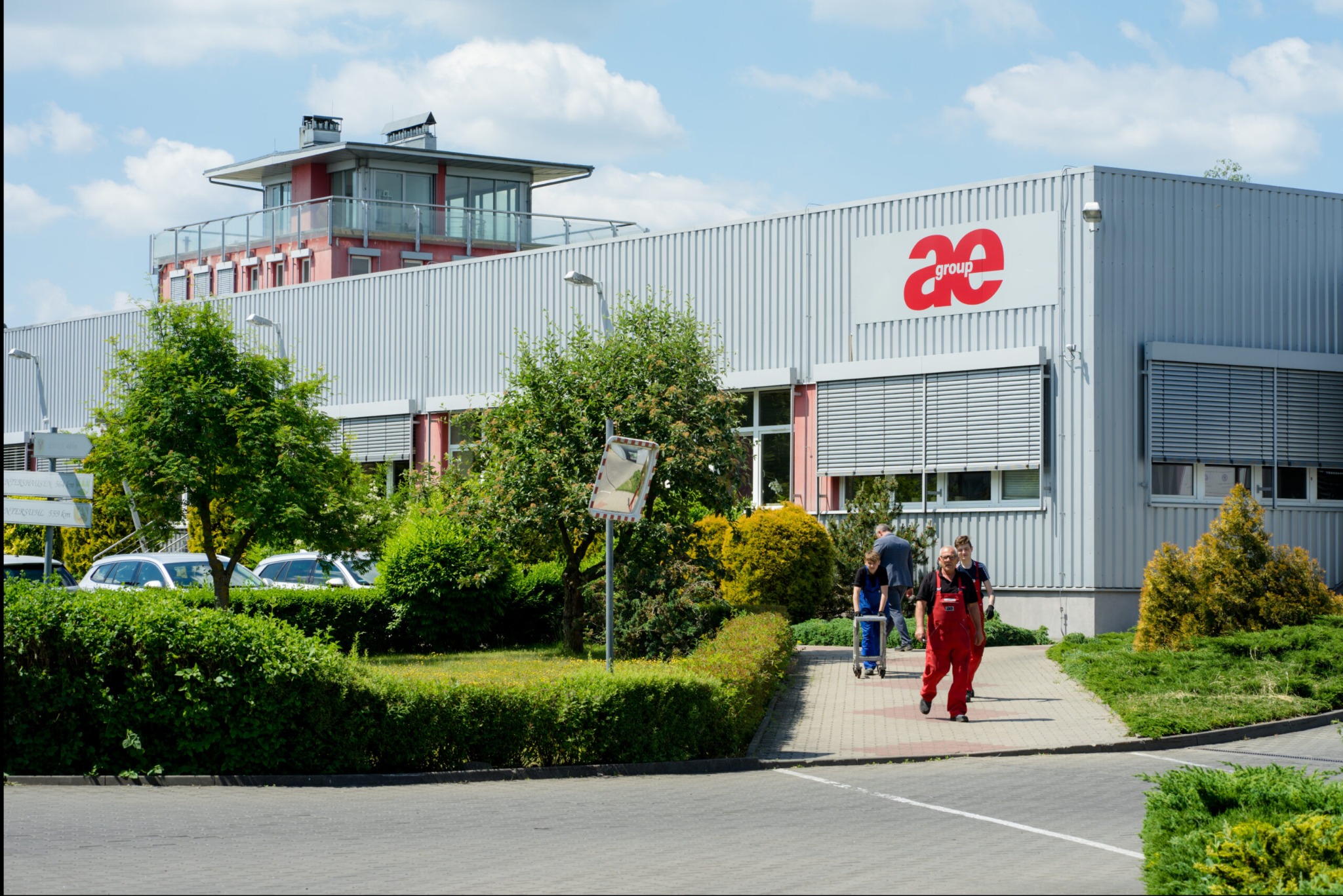 Budynek firmy AE Group w słoneczny dzień. Pracownicy w odzieży roboczej wchodzą do budynku. Widoczne logo firmy na fasadzie i zadbana zieleń wokół.