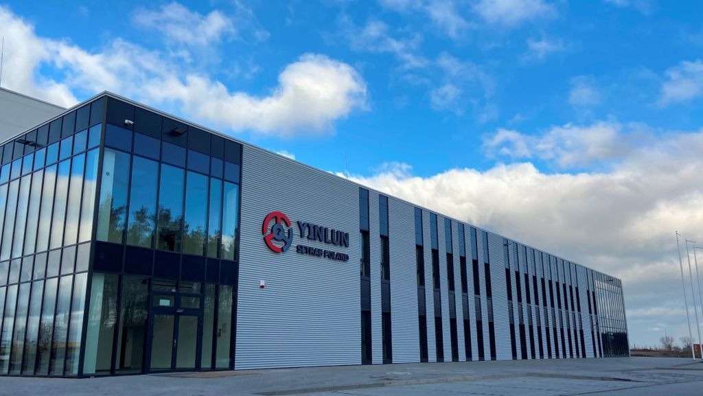 Nowoczesny budynek biurowy z szarymi panelami i dużymi oknami, logo firmy Yinlun Setrab Poland, błękitne niebo z chmurami w tle. Architektura komercyjna.