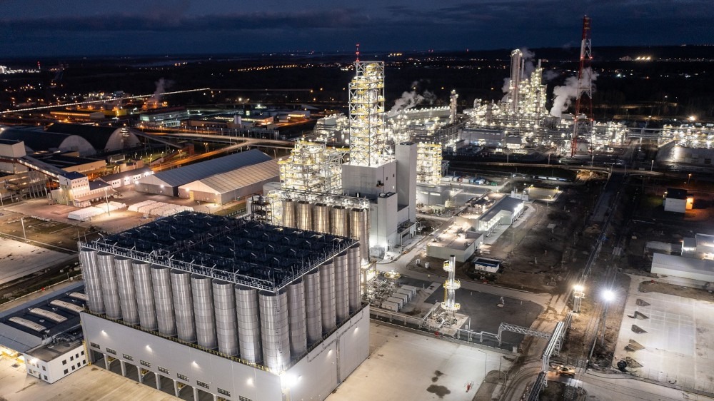 Nocny widok z lotu ptaka na rozległy, oświetlony kompleks przemysłowy z licznymi zbiornikami i wieżami, z delikatną poświatą miejską w tle.
