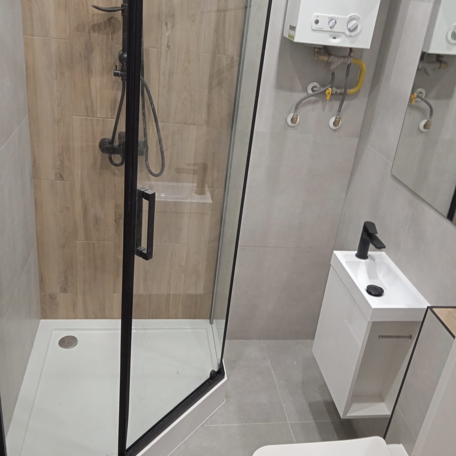 Nowoczesna łazienka z kabiną prysznicową z czarnymi profilami, armaturą w kolorze czarnym, umywalką nablatową na szafce i płytkami imitującymi drewno na ścianie prysznica.