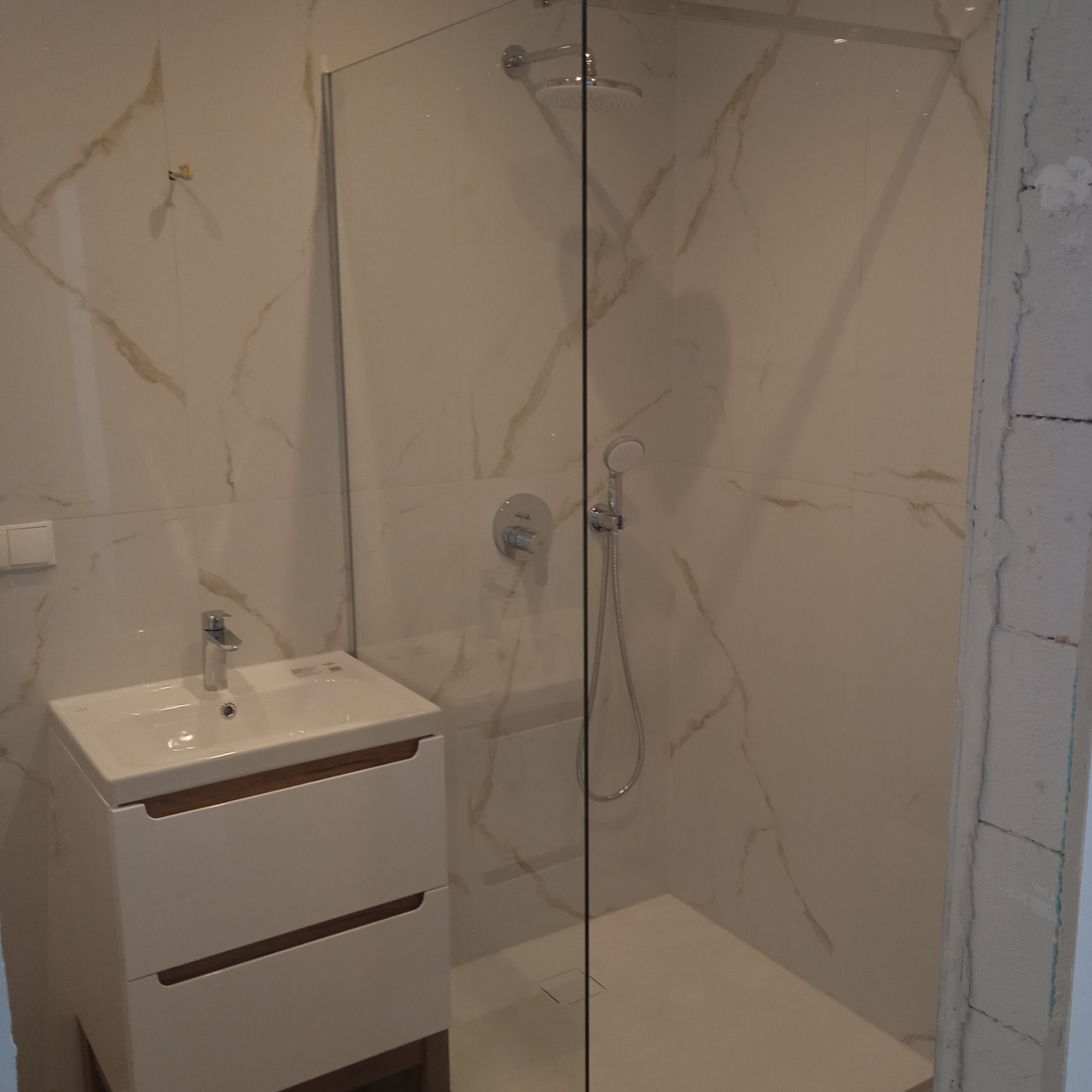 Biała umywalka z szafką i kabina prysznicowa z deszczownicą na tle marmurowych płytek w wykończonej łazience. Widoczny fragment surowej ściany.