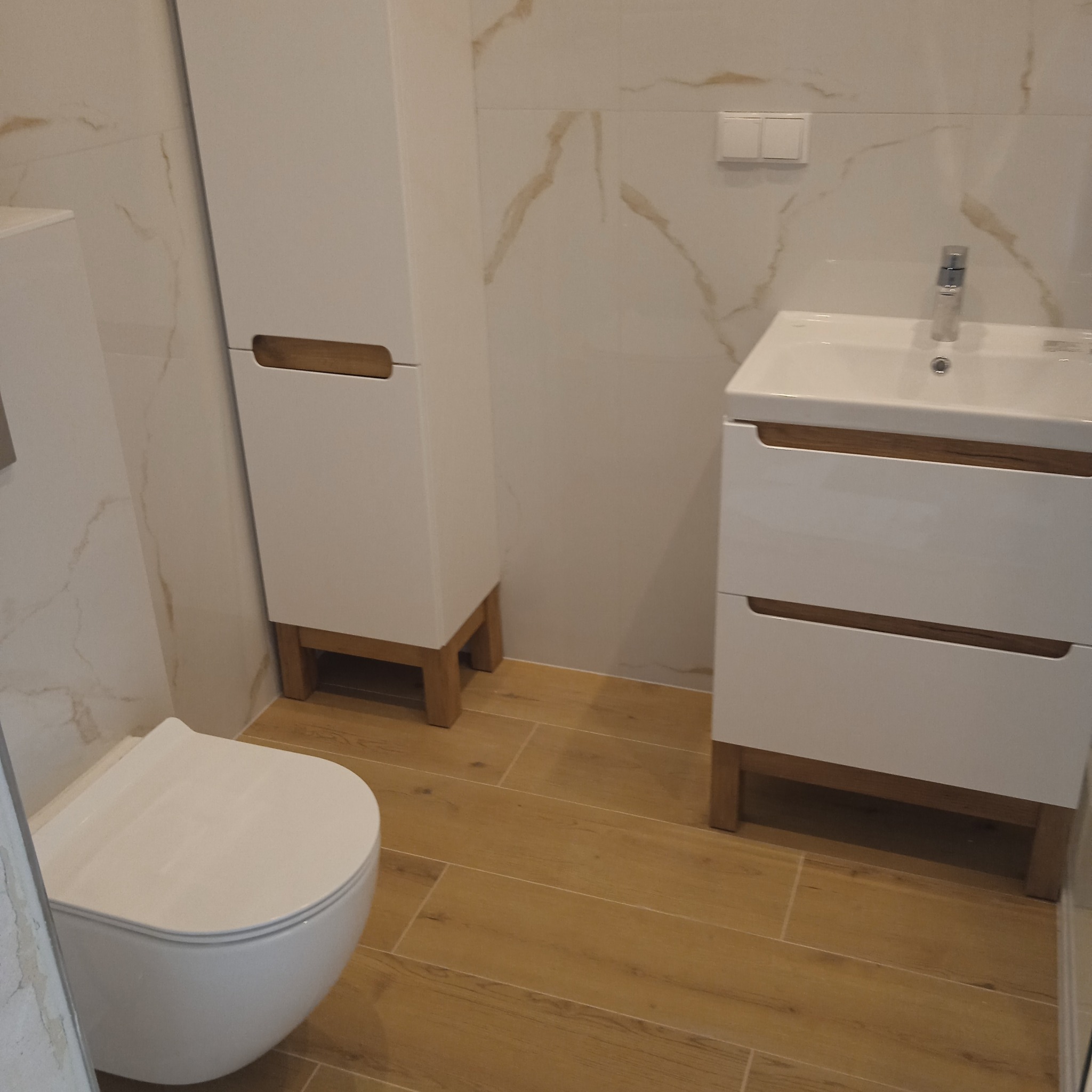 Nowoczesna łazienka po remoncie: białe meble z drewnianymi akcentami, marmurowe płytki ścienne i podłoga imitująca drewno. Widoczna toaleta, szafka z umywalką i słupek.