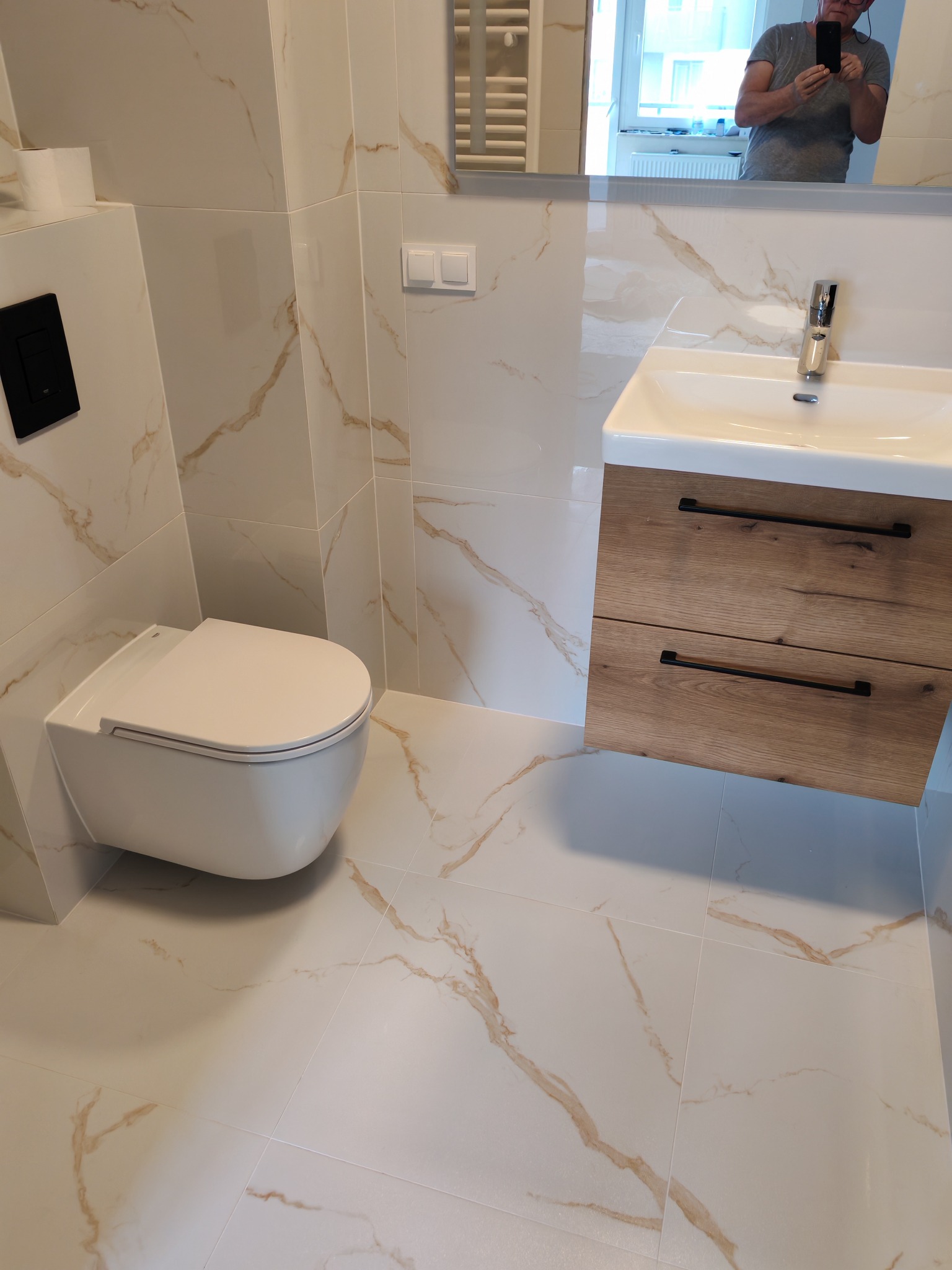 Nowoczesna łazienka z wiszącą toaletą i umywalką z drewnianą szafką. Płytki imitujące marmur na ścianach i podłodze. W lustrze odbija się osoba robiąca zdjęcie.