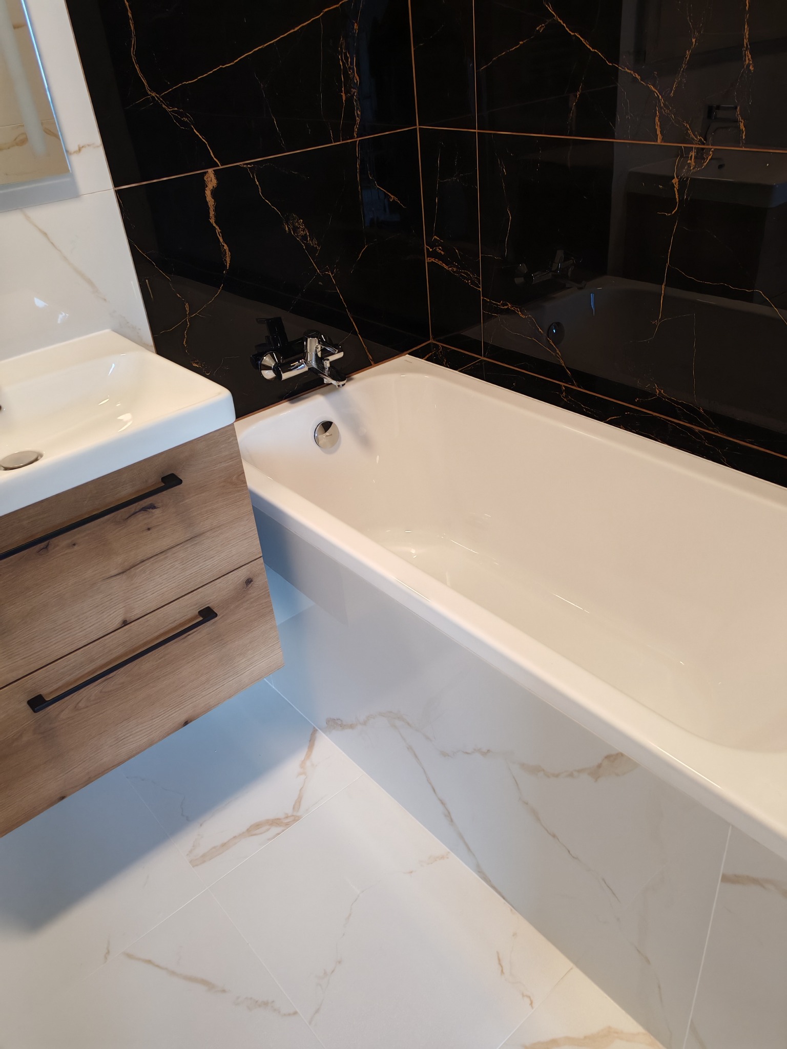 Nowoczesna łazienka: biała wanna, umywalka z drewnianą szafką i czarne płytki z marmurowym wzorem. Minimalistyczny design z akcentem luksusu.