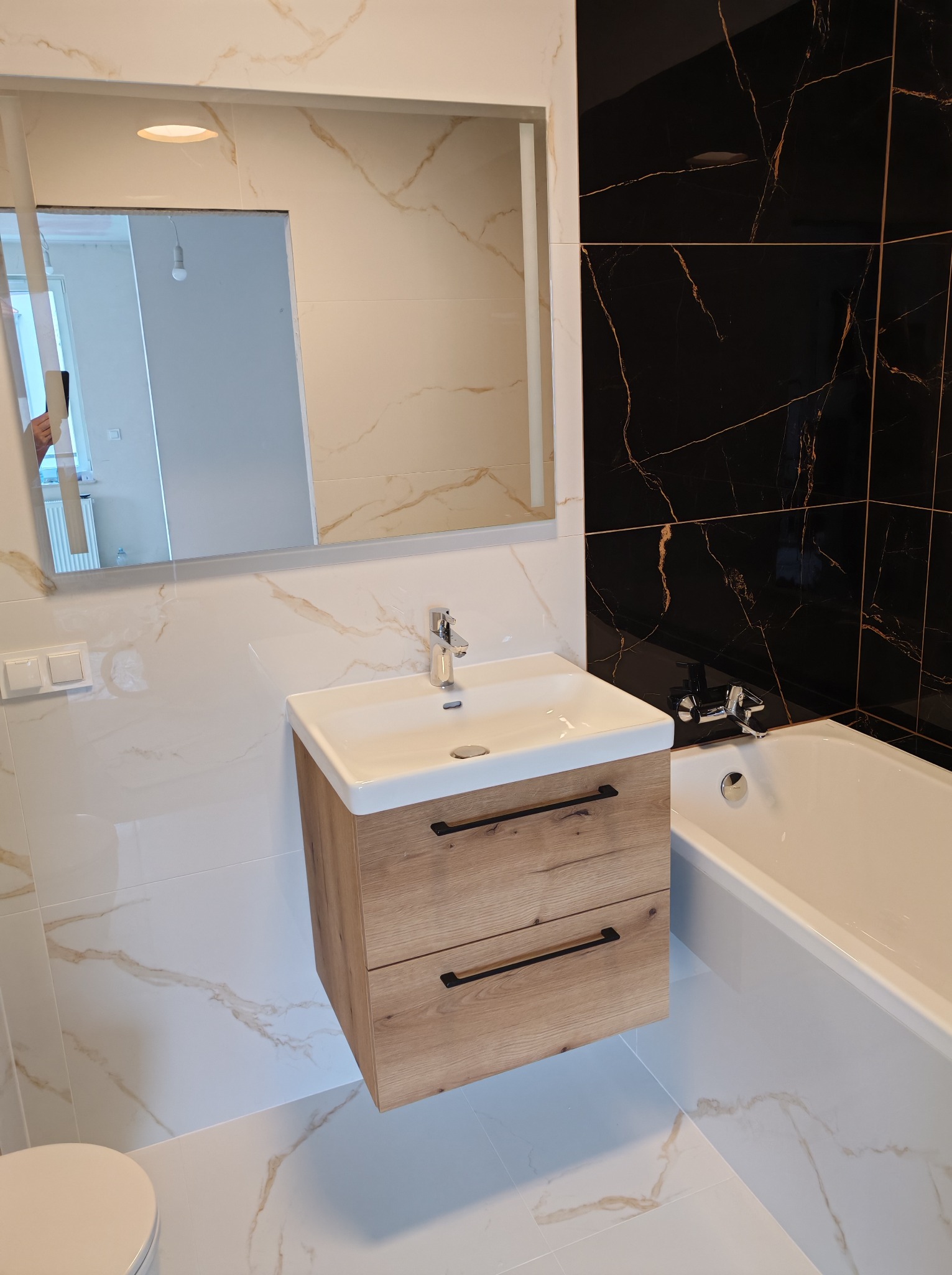 Nowoczesna łazienka po remoncie: białe i czarne płytki z marmurowym wzorem, drewniana szafka pod umywalką, lustro i wanna. Minimalistyczny design z akcentami złota.