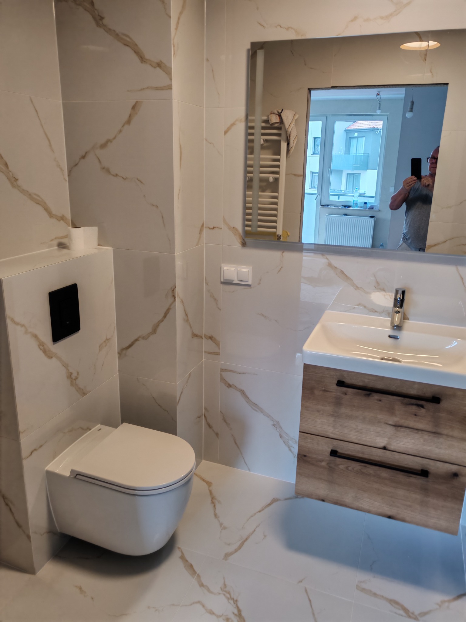 Nowoczesna łazienka z białymi płytkami imitującymi marmur, podwieszana toaleta i szafka z umywalką z drewnianymi frontami. W lustrze odbija się osoba robiąca zdjęcie.