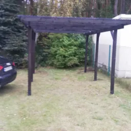 Ciemna, drewniana wiata garażowa na trawniku, z samochodem w tle i drzewami w tle.