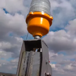 Pomarańczowa lampa ostrzegawcza lotnicza zamontowana na metalowej konstrukcji, widok z dołu na tle pochmurnego nieba i zabudowań miejskich w oddali.