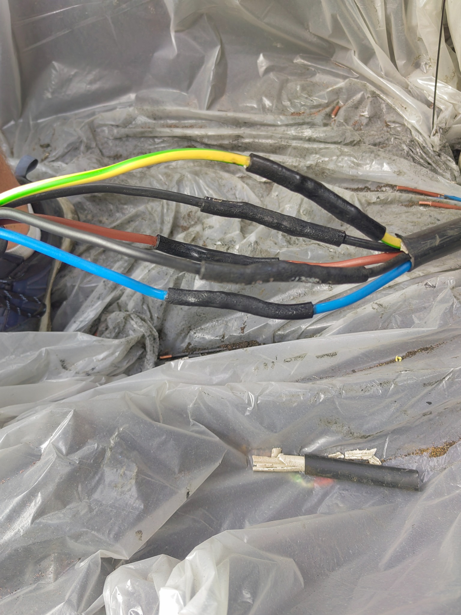 Przygotowane kable elektryczne z kolorowymi żyłami, zabezpieczone termokurczliwą izolacją, leżące na folii ochronnej. Fragment niebieskiego buta w lewym górnym rogu kadru.