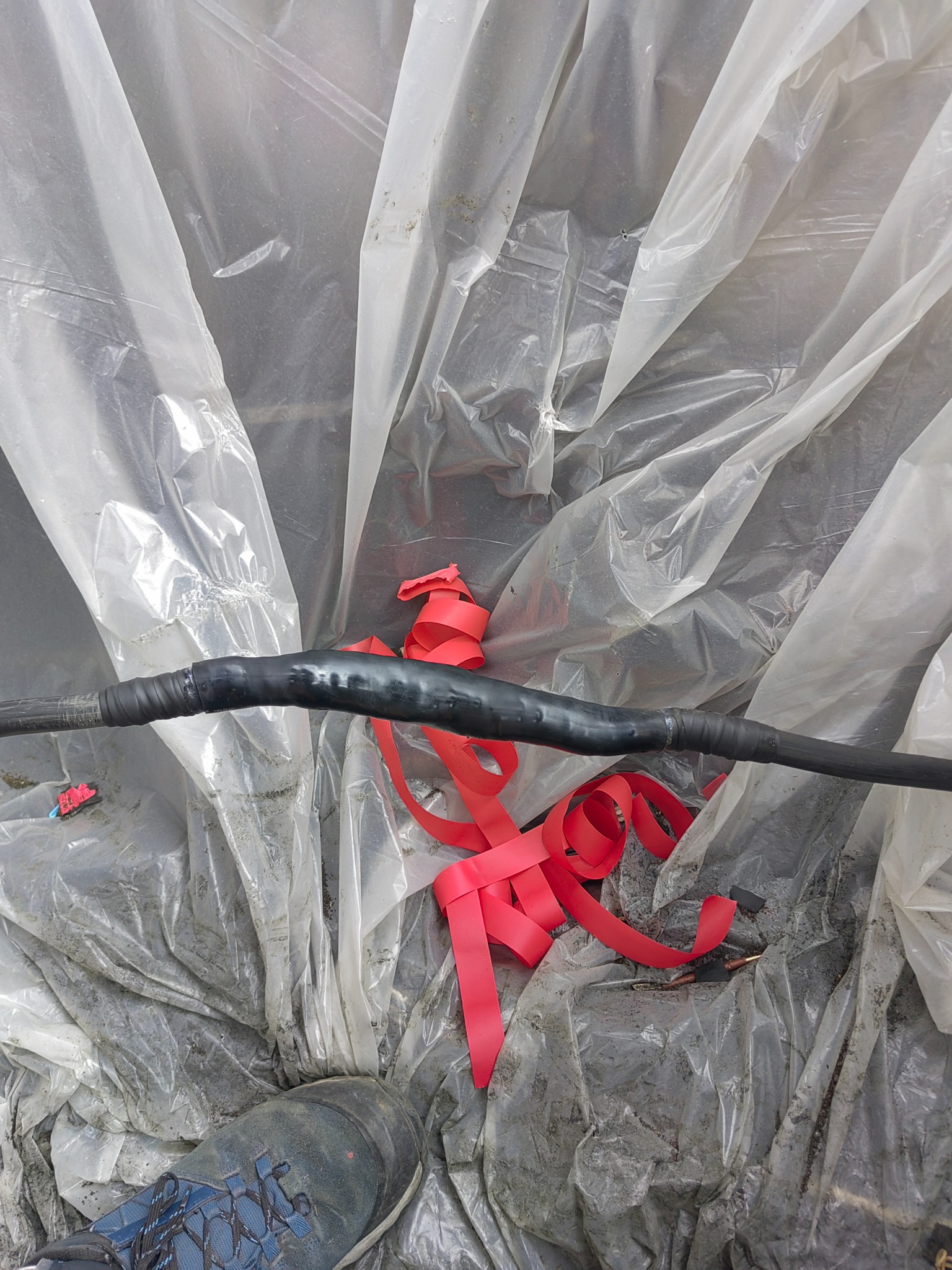 Połączenie kabla elektrycznego zaizolowane czarną taśmą, widoczne czerwone paski taśmy izolacyjnej. W tle folia budowlana i fragment buta roboczego.