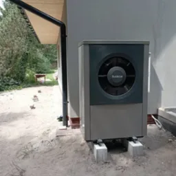 Szara jednostka zewnętrzna z logo Buderus, zamontowana na betonowych blokach przy szarej ścianie budynku z drewnianą podbitką i rynną, na piaszczystym podłożu.