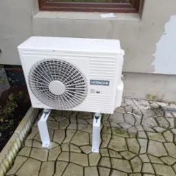 Biała jednostka zewnętrzna klimatyzacji Hitachi zamontowana na metalowych wspornikach przed budynkiem z nierówną elewacją, widoczne krople wody na obudowie, kostka brukowa i fragment ogrodu.