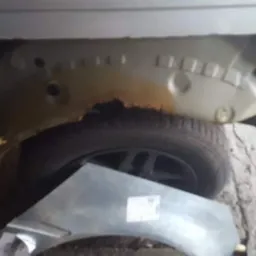 Fragment nadkola samochodu z widoczną korozją, obok leży nowy element blacharski przed montażem.