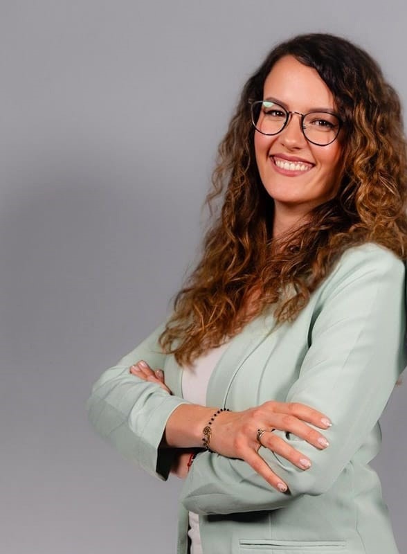 Uśmiechnięta kobieta w okularach i pastelowej marynarce, z założonymi rękami, prezentująca się na szarym tle.