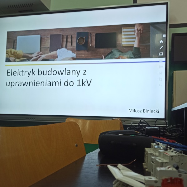 Sala szkoleniowa z wyświetlanym na ekranie tytułem 'Elektryk budowlany z uprawnieniami do 1kV' oraz rozłożonymi na stole elementami elektrycznymi i dwoma krzesłami w pierwszym planie.