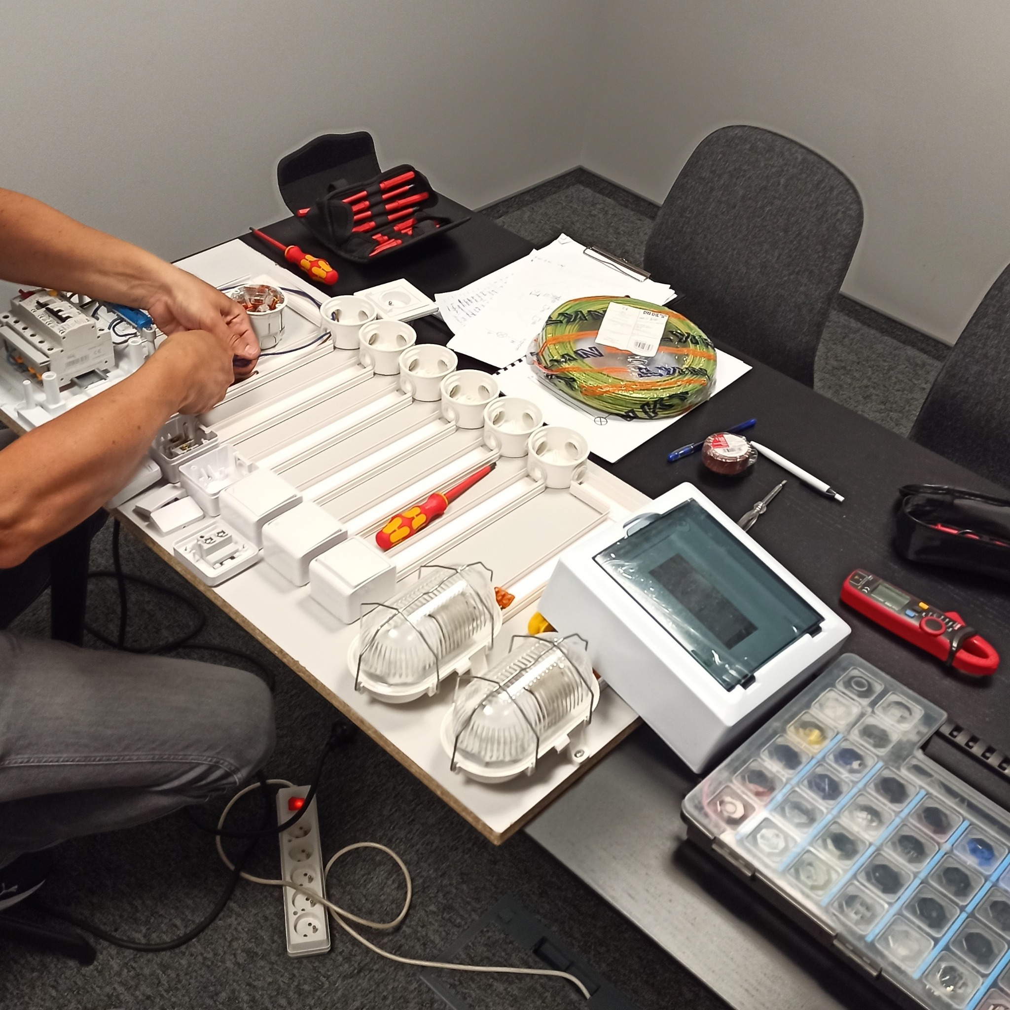 Osoba podłącza przewody do gniazdka na makiecie instalacji elektrycznej podczas szkolenia, widoczne narzędzia, przewody, elementy instalacji i gniazdka, na drugim planie zwój kabli i dokumentacja.